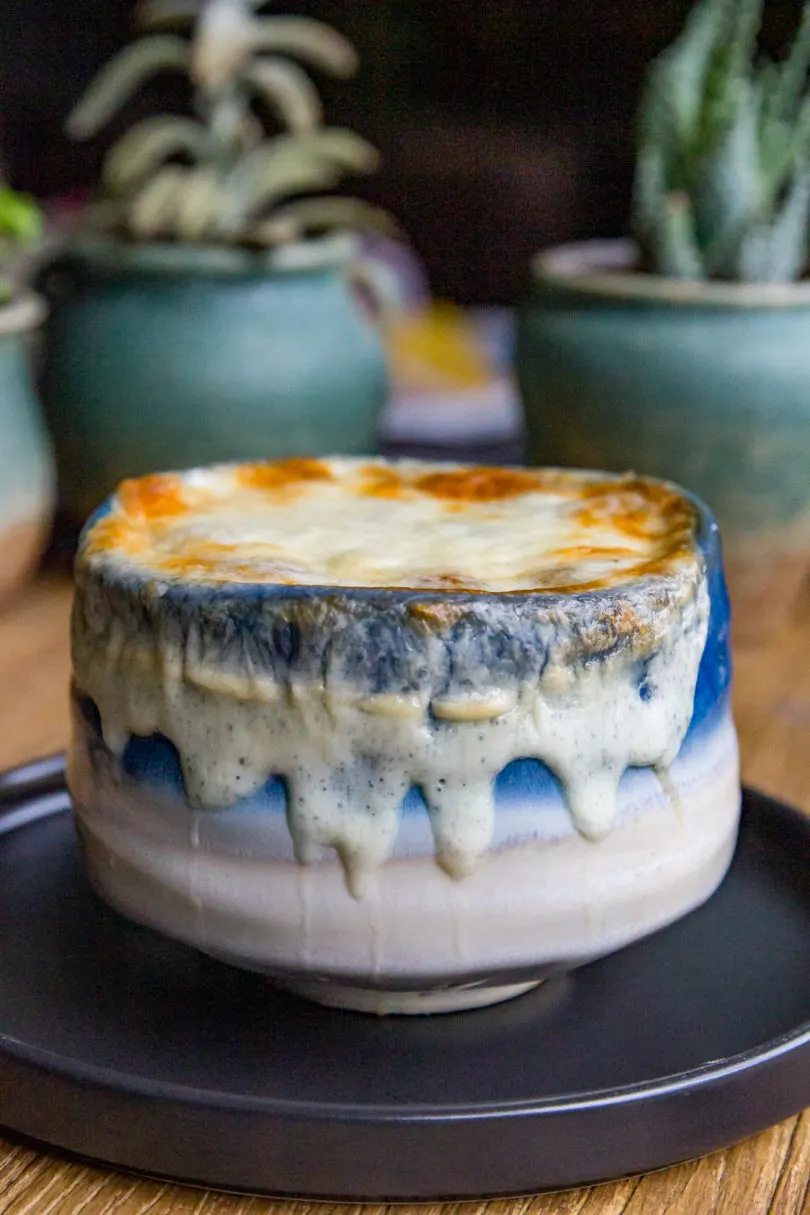 Blue ombre bowl of Smoked French Onion Soup with melted white provolone cheese that's been browned lightly around the top edges. On a black plate.