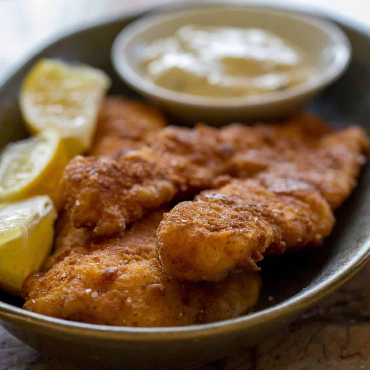 Crispy fried fish fillets served with lemon wedges and tartar sauce on a black plate.