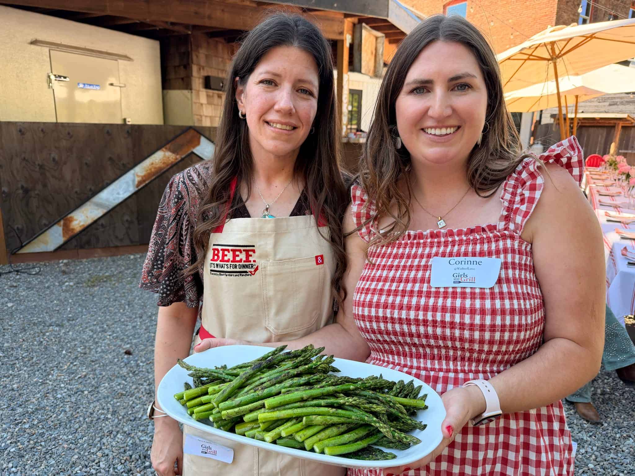 Two women smiling, holding a tray of grilled asparagus outdoors.
