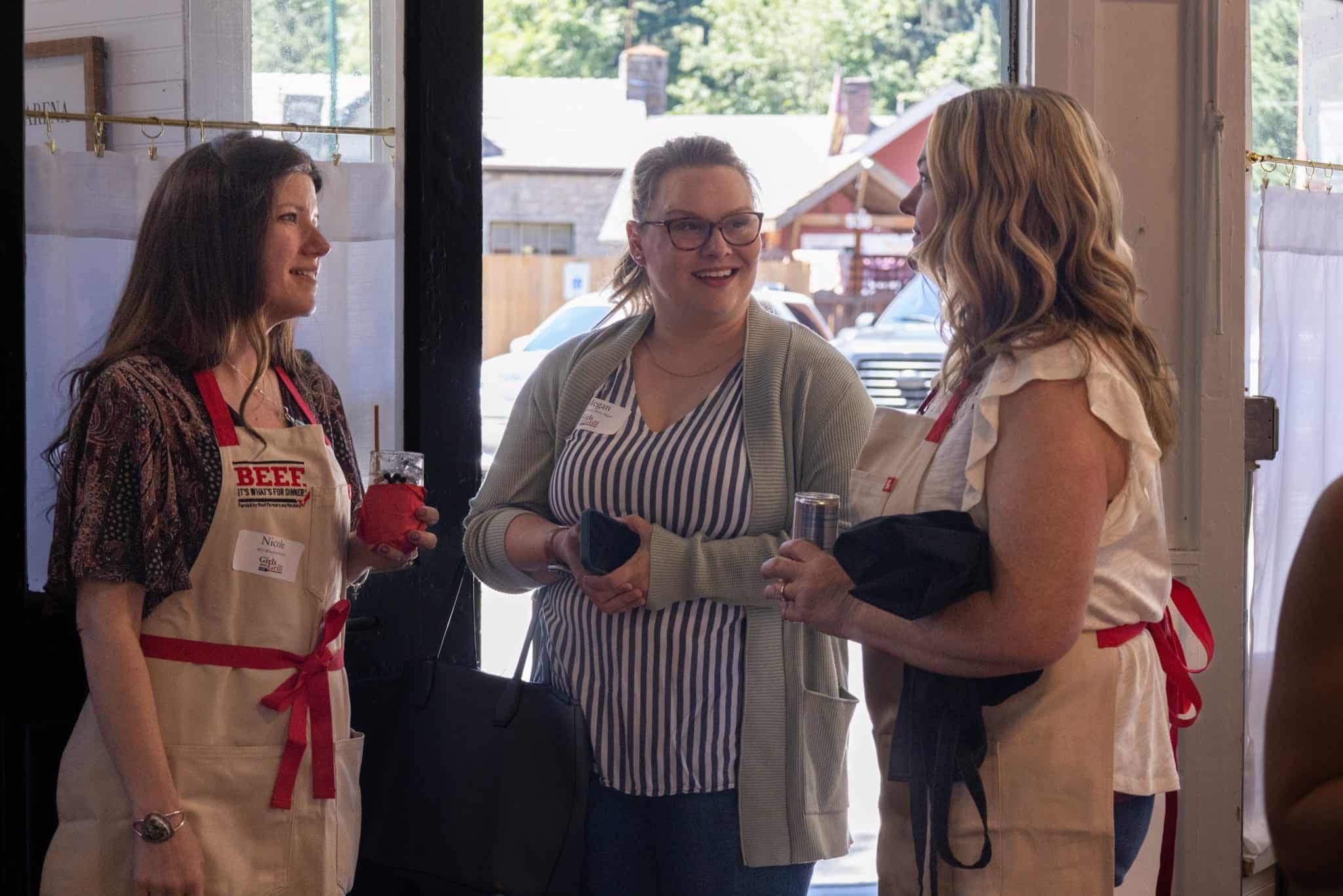 Three women in aprons chatting at a barbecue event.