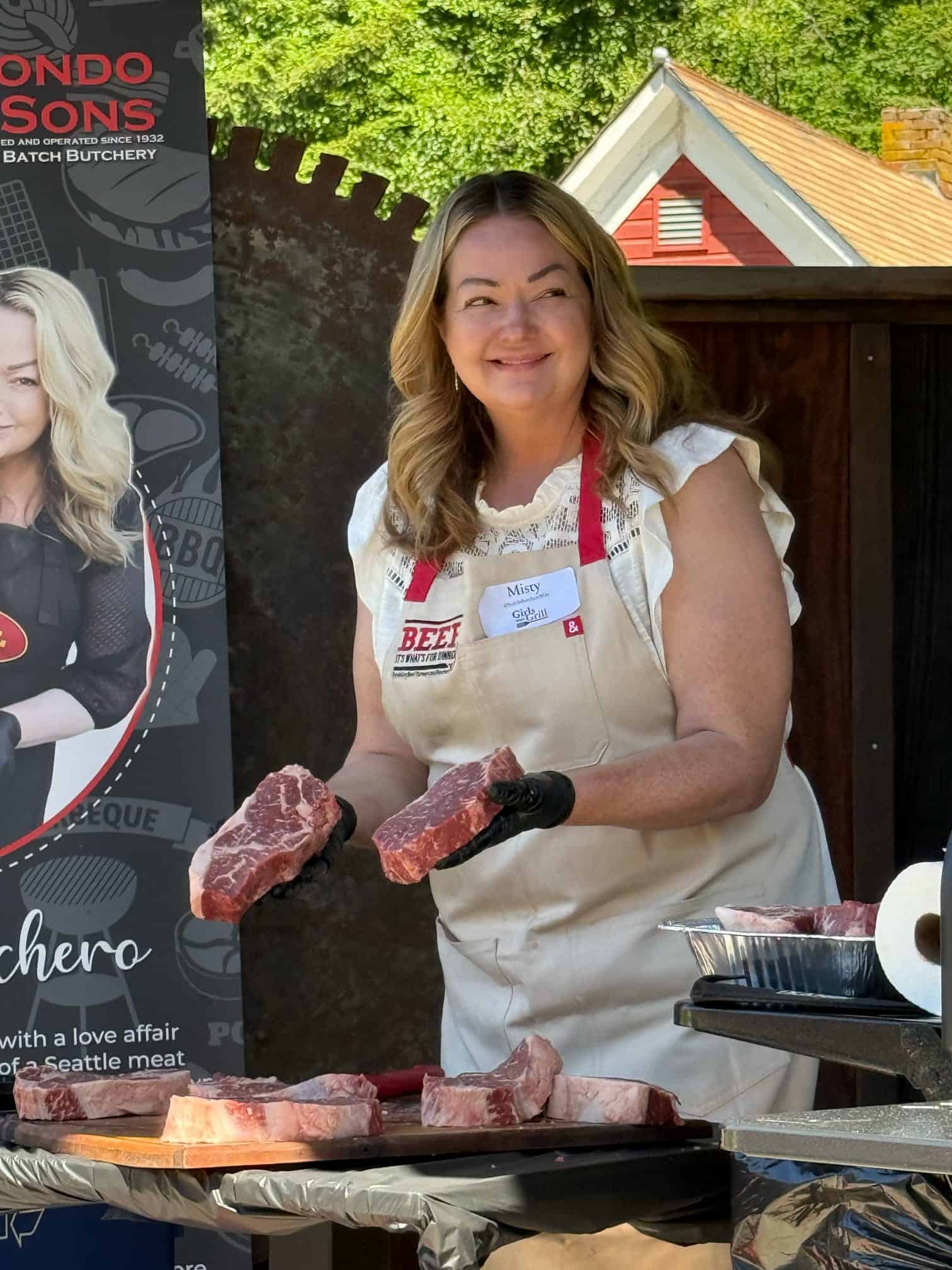 Young woman smiling while holding raw steak pieces at outdoor barbecue.