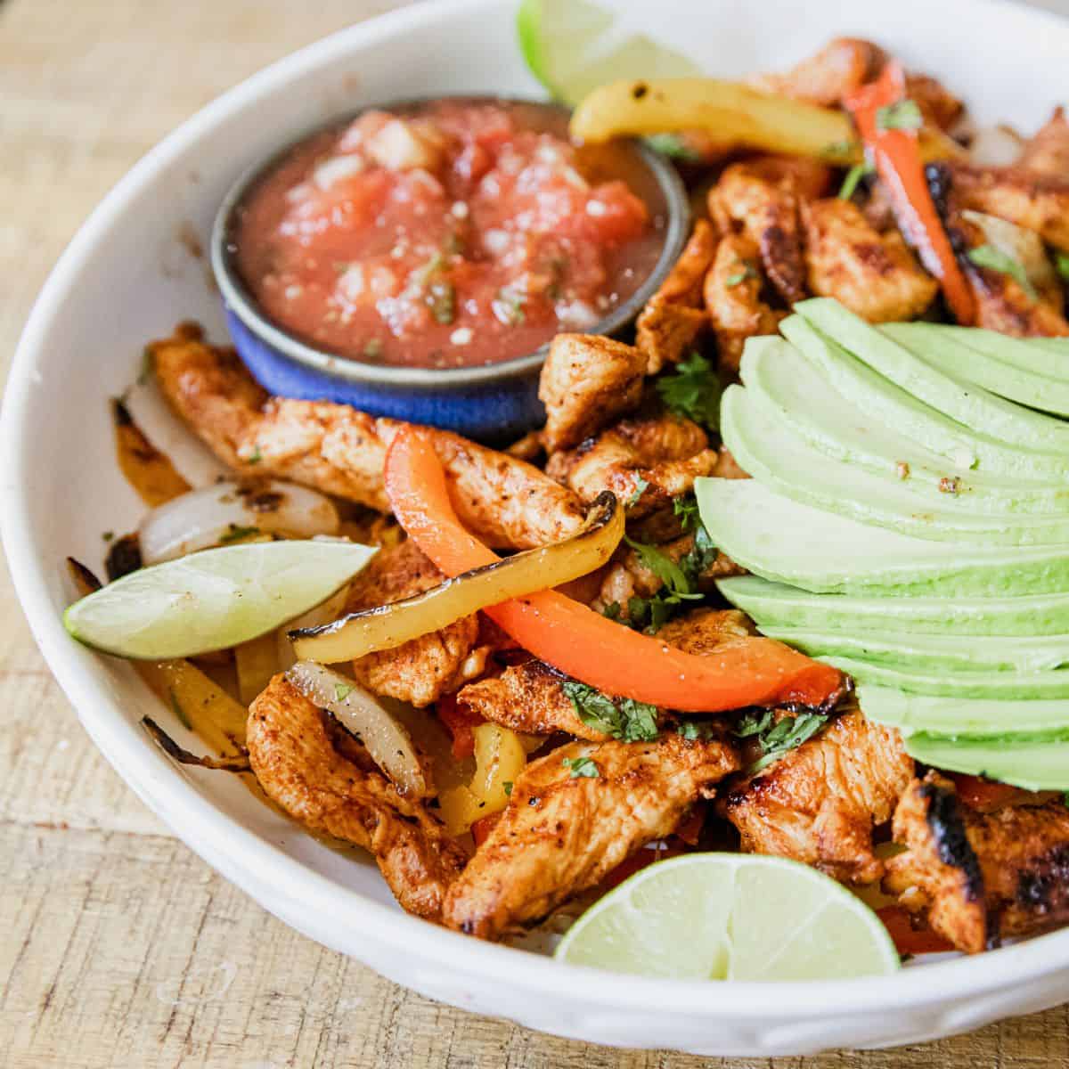 Delicious grilled chicken fajitas with fresh avocado slices and salsa in a white bowl.