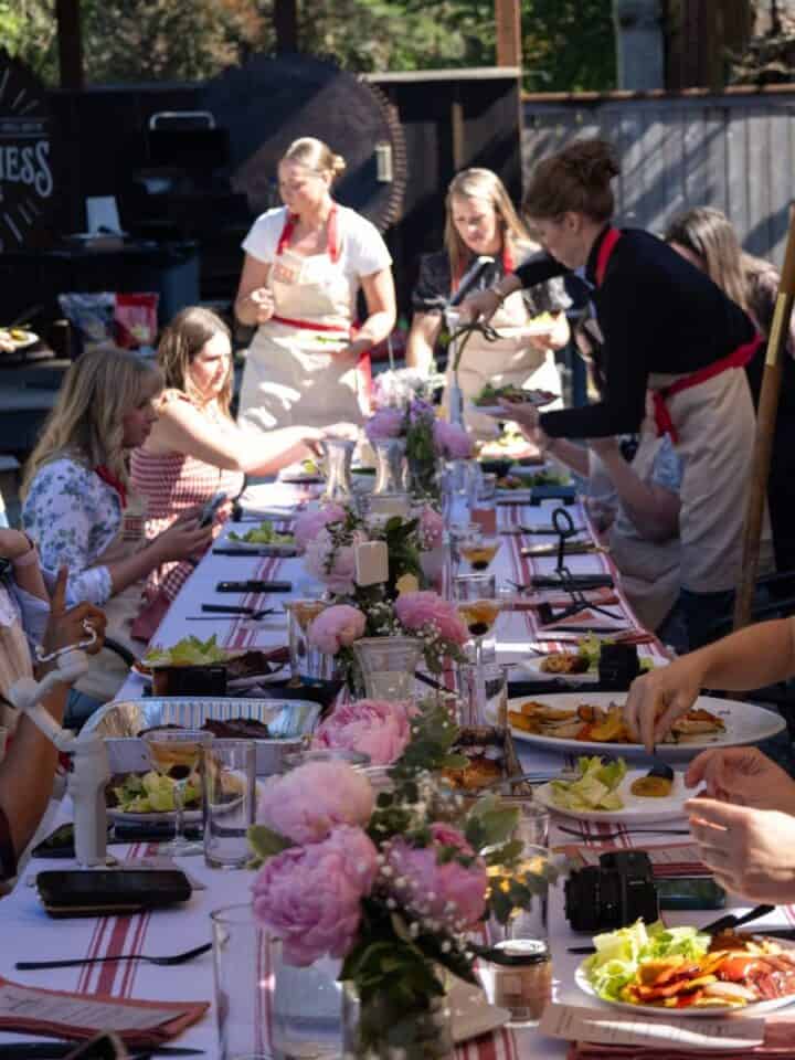 Group of girls and women enjoying outdoor dinner with grilled food and flowers.