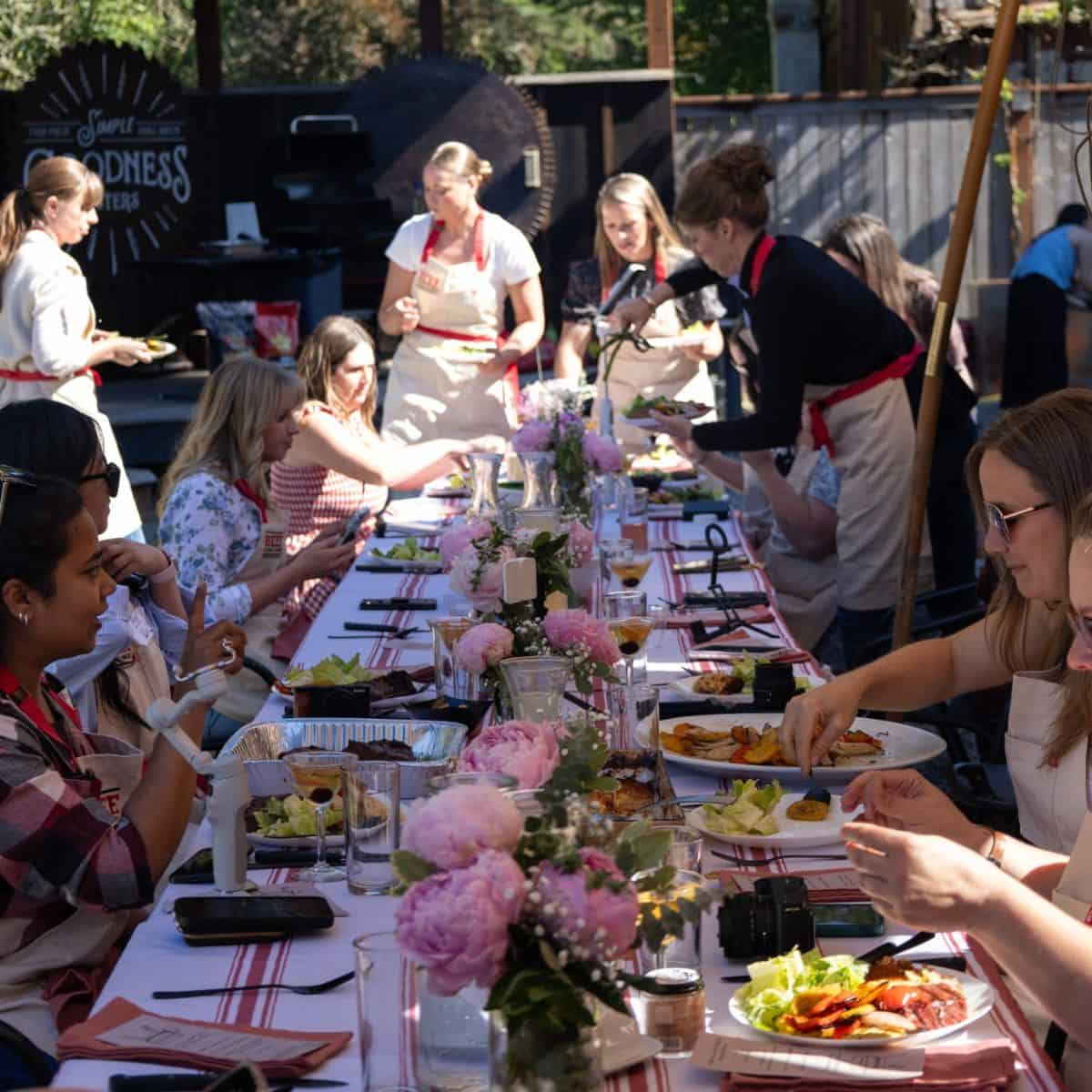 Group of girls and women enjoying outdoor dinner with grilled food and flowers.