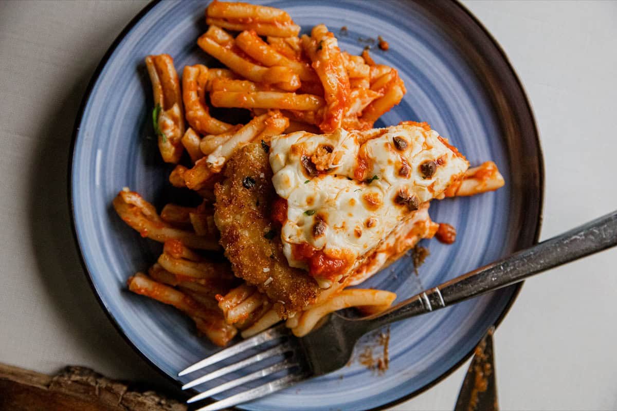 Chicken Parmesan with pasta on a blue plate, served with a fork.
