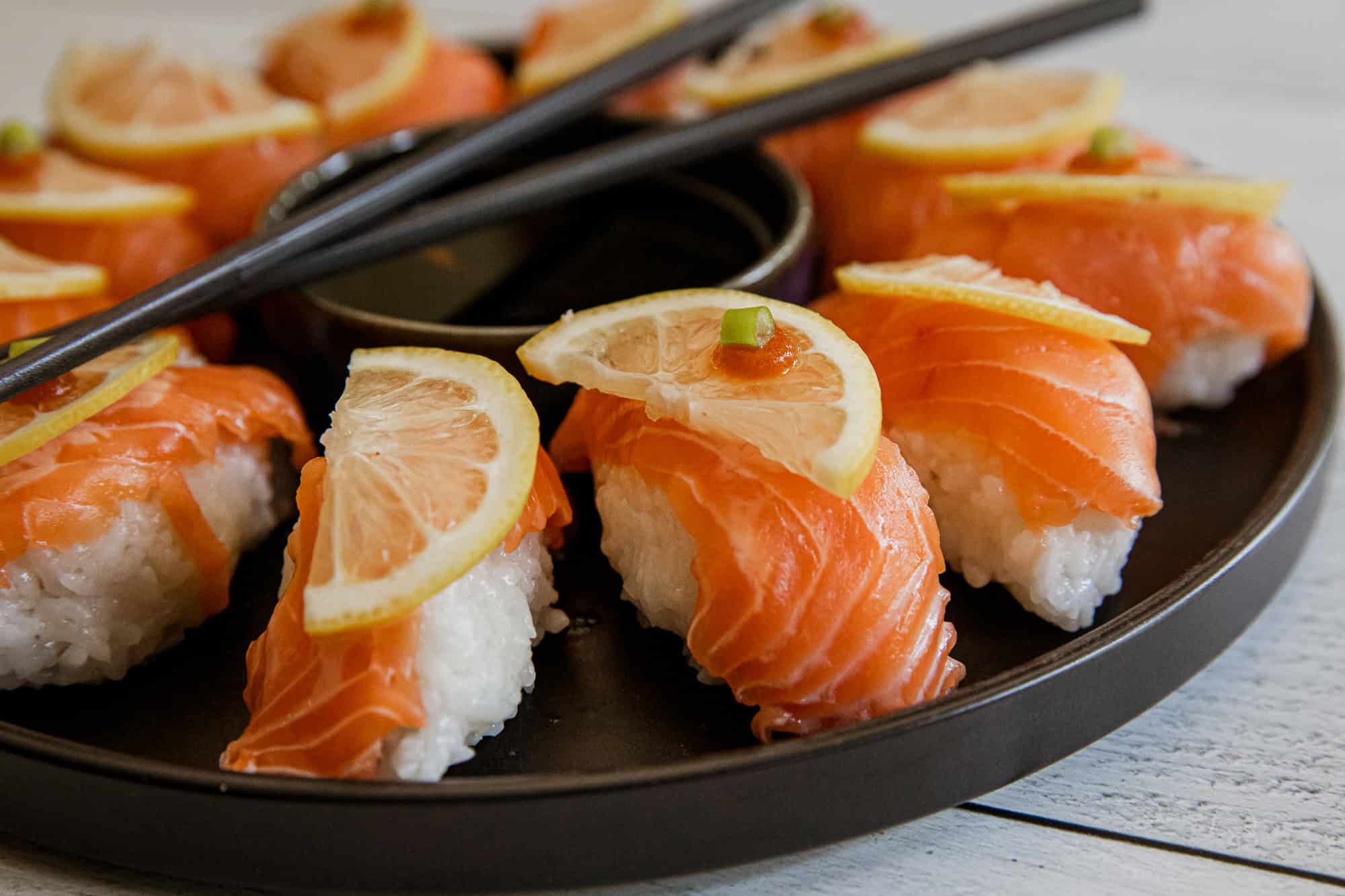 Salmon nigiri sushi with lemon slices on top, served on a black plate.