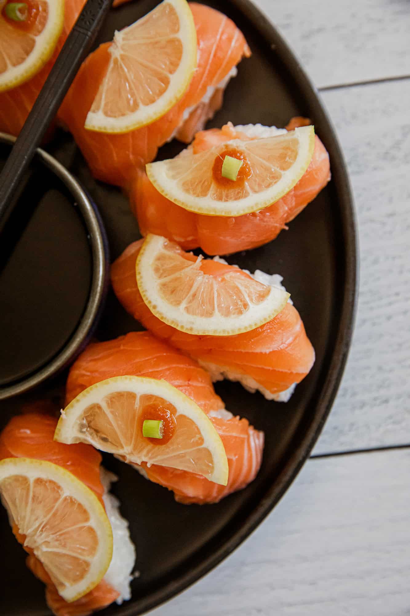 Fresh salmon nigiri topped with lemon slices and green onion on a black plate.
