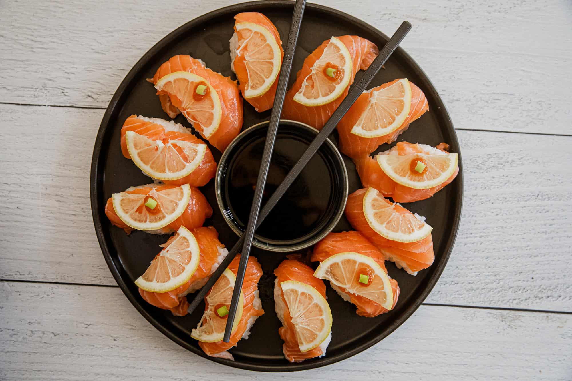 Salmon nigiri sushi with lemon slices and soy sauce in a black dish.