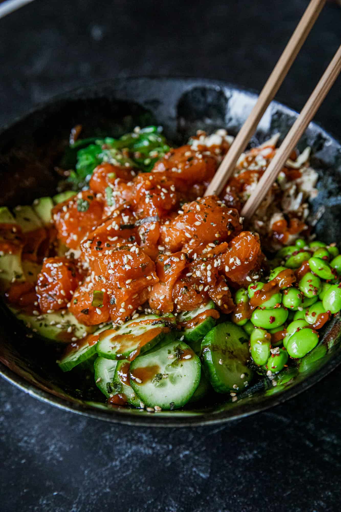 Close-up of spicy salmon, cucumber, and edamame in a black bowl with chopsticks.