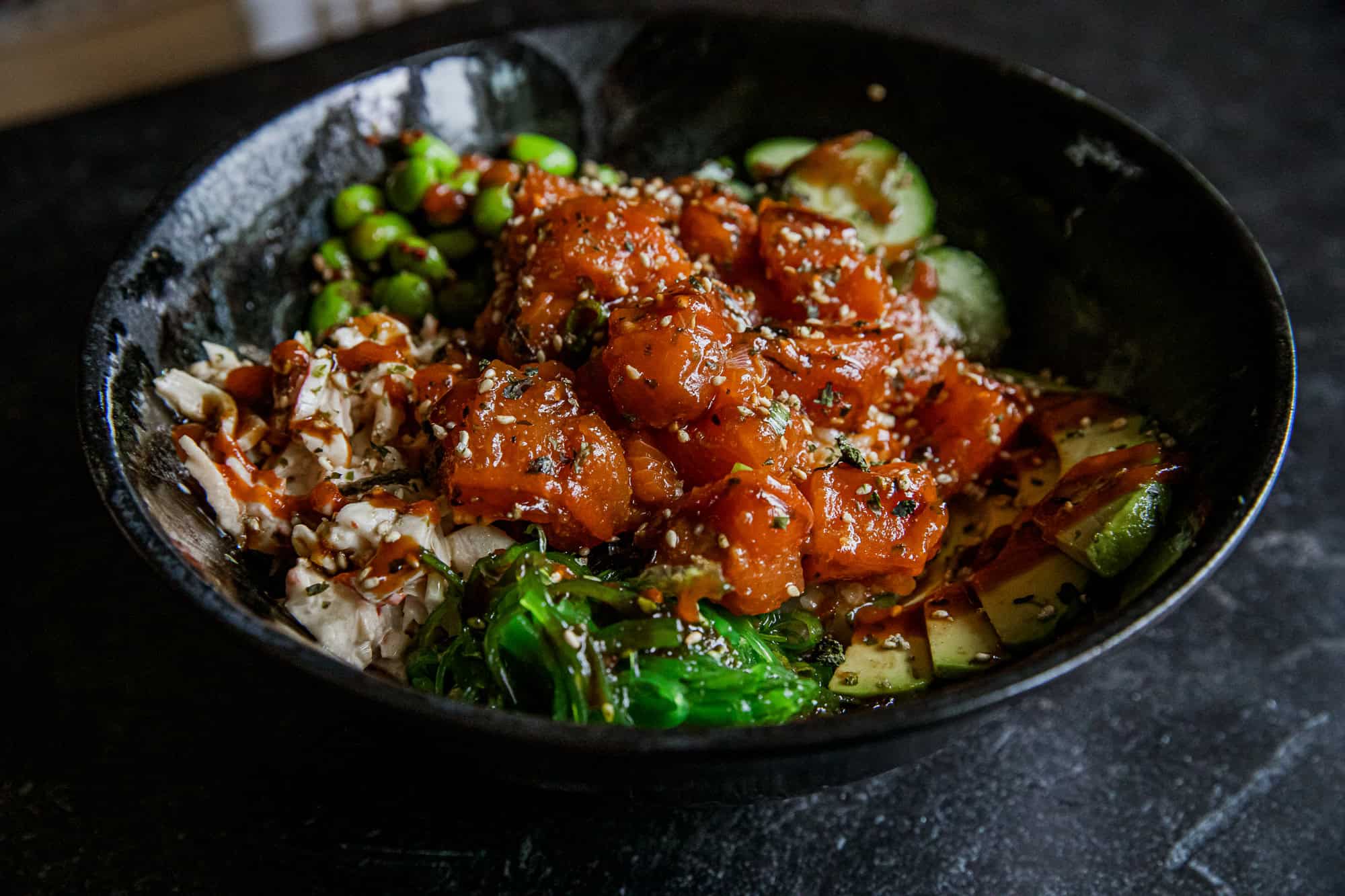 Close-up of spicy salmon poke bowl with vegetables and rice in black bowl.