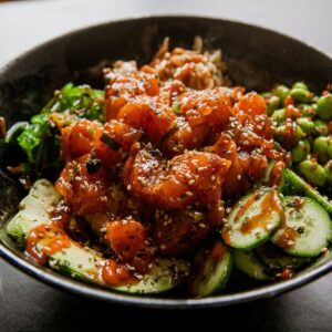 Spicy salmon poke bowl with cucumbers and sesame seeds in a black bowl.