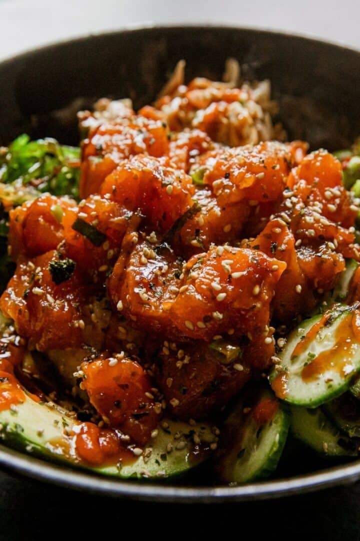 Spicy salmon poke bowl with cucumbers and sesame seeds in a black bowl.