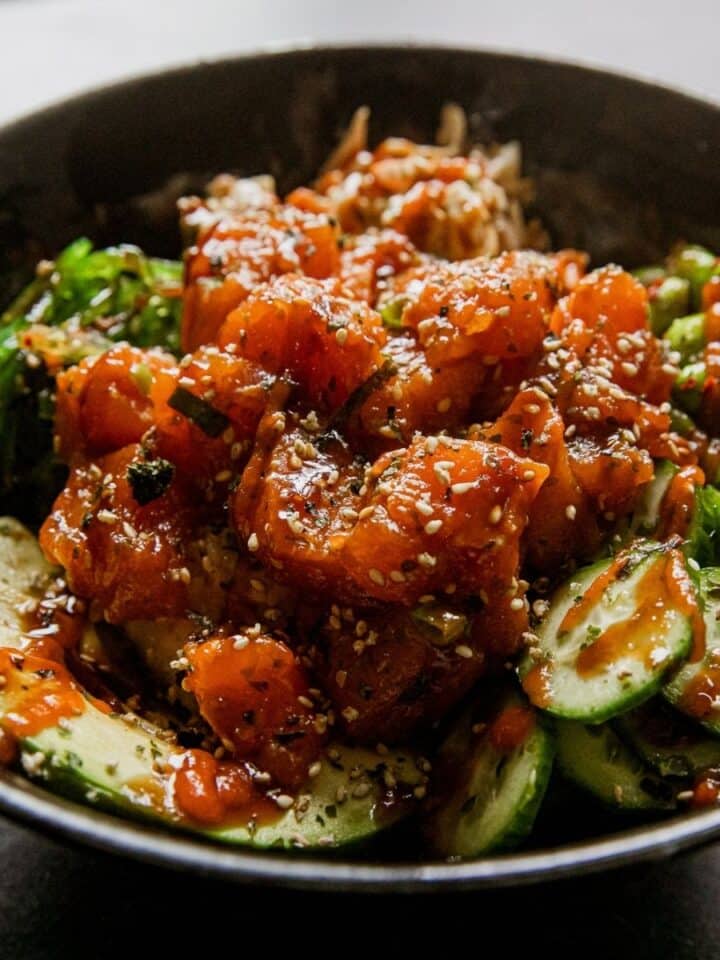 Spicy salmon poke bowl with cucumbers and sesame seeds in a black bowl.