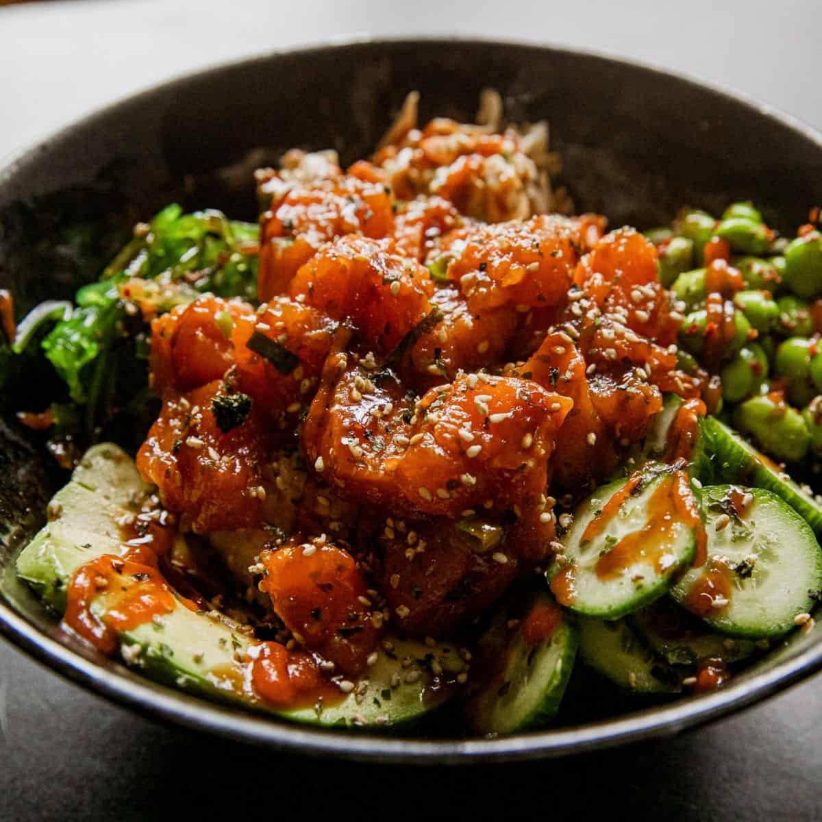 Spicy salmon poke bowl with cucumbers and sesame seeds in a black bowl.