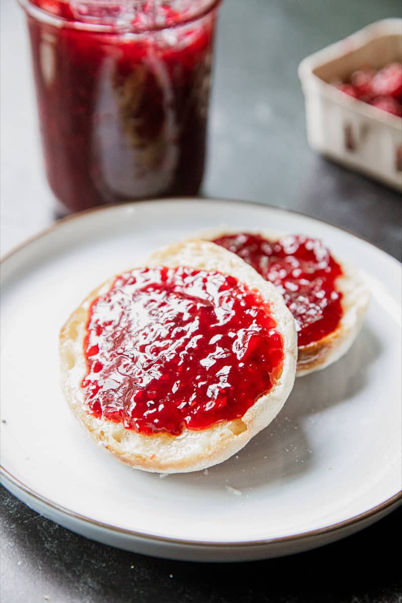 Two bread slices with tayberry jam on a white plate, jar of jam in background.