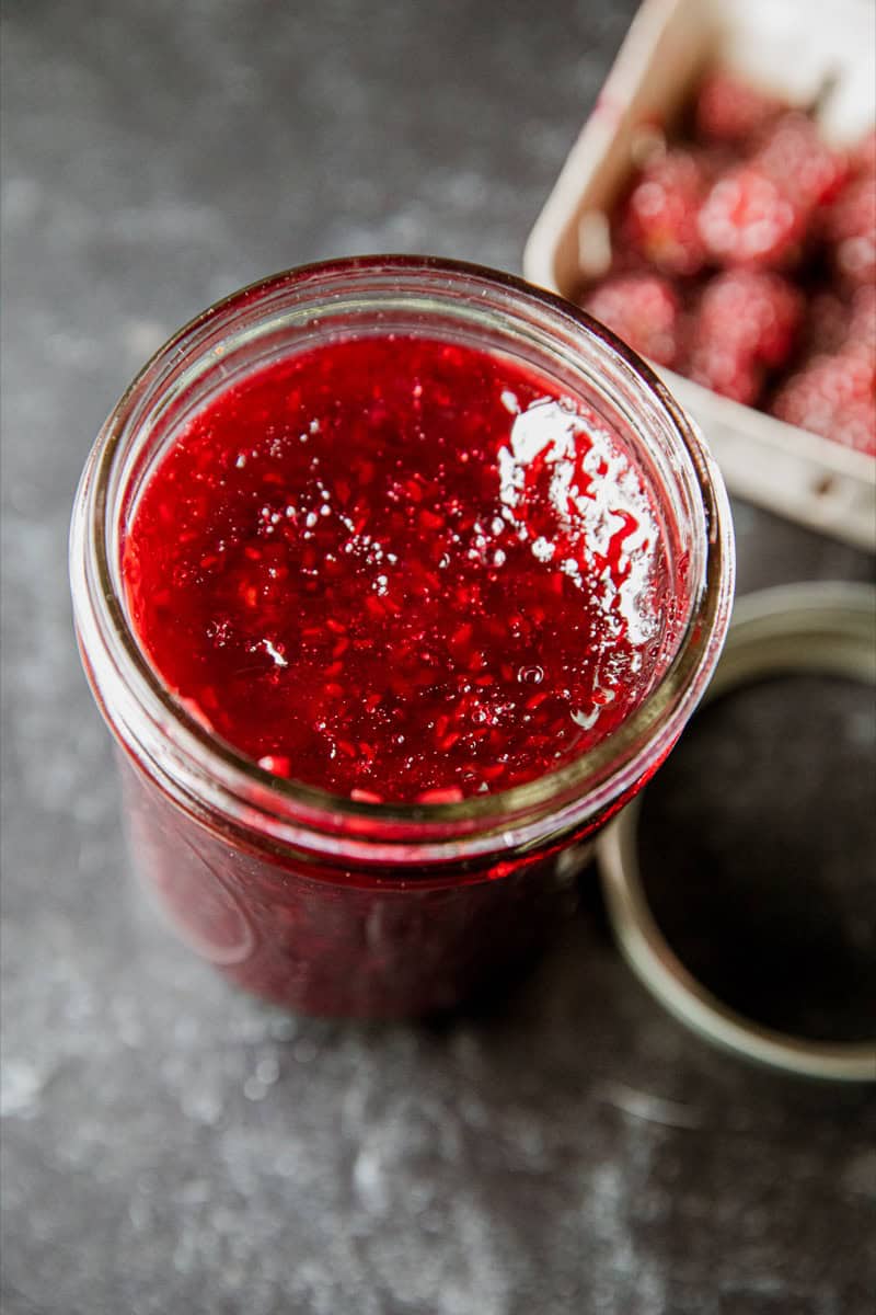 Close-up of fresh tayberry jam in a glass jar with a basket of tayberries in the background.