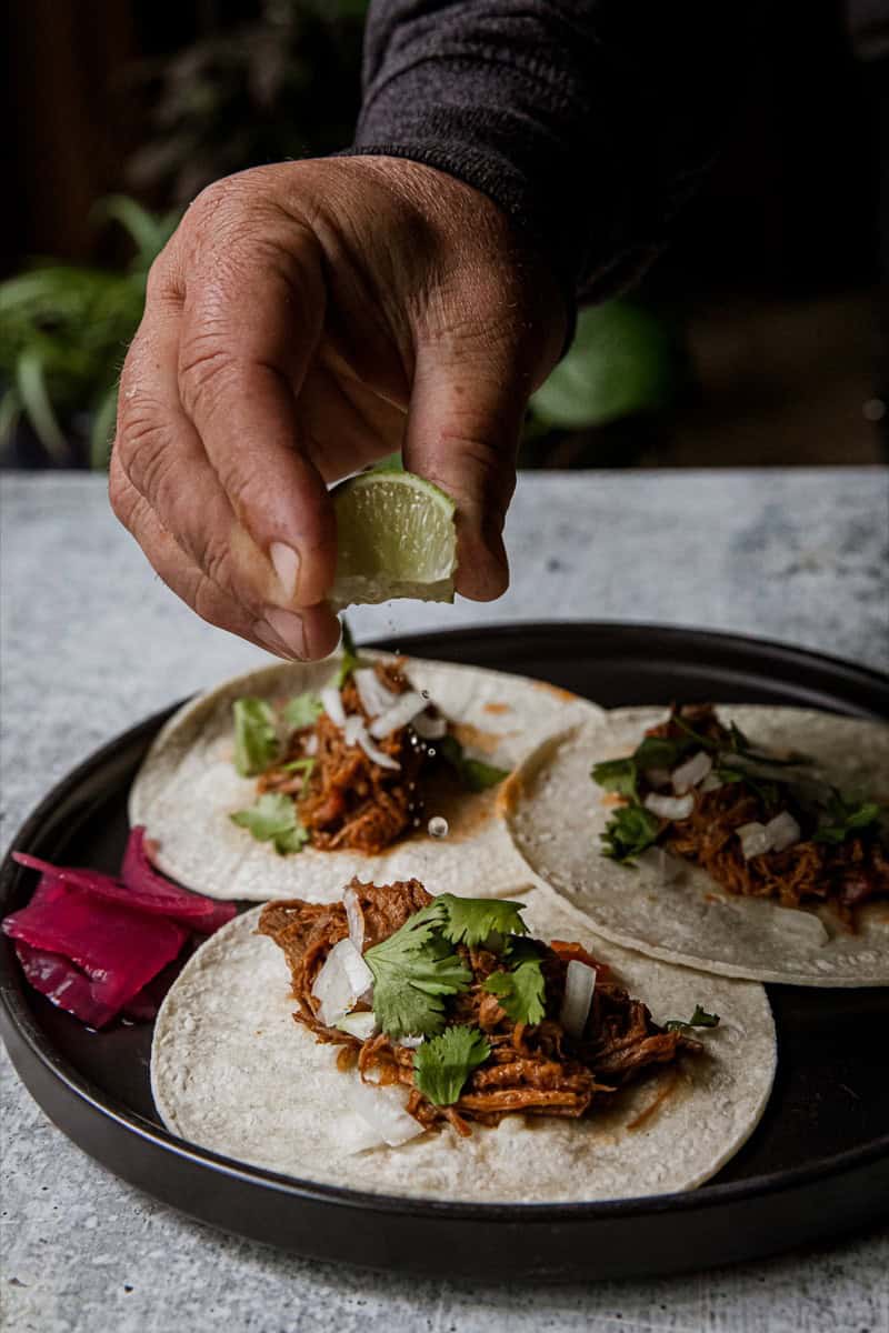 Close-up of hand squeezing lime over venison barbacoa tacos on black plate.