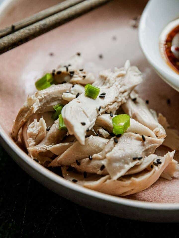 Steamed chicken slices garnished with scallions and black sesame seeds on a pink plate.