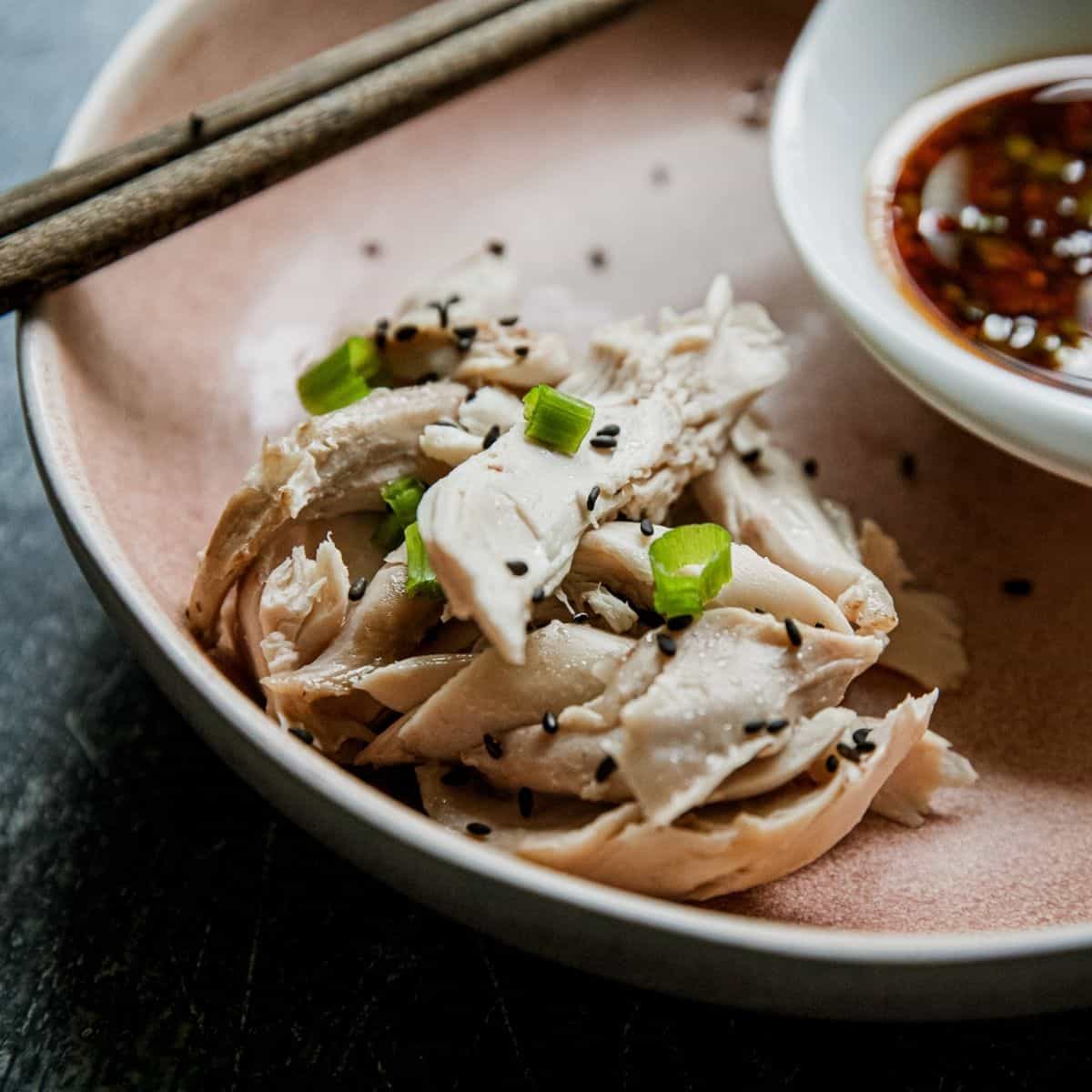 Steamed chicken slices garnished with scallions and black sesame seeds on a pink plate.