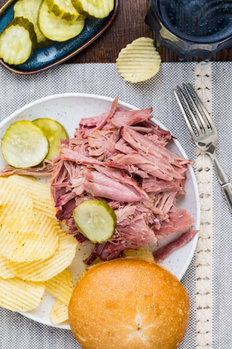 Juicy double smoked picnic ham with pickles and potato chips on a white plate.
